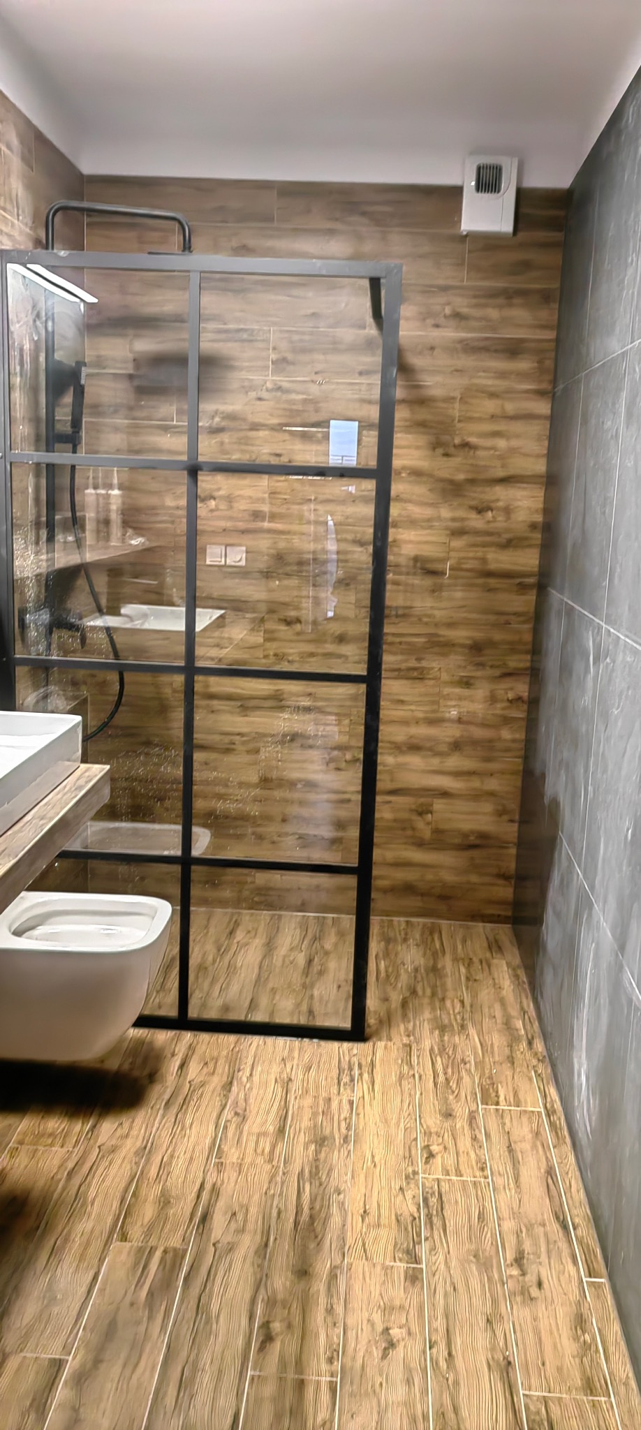 Nowoczesna łazienka z kabiną prysznicową typu walk-in z czarnymi profilami, wiszącą toaletą i drewnopodobnymi płytkami na podłodze i ścianie. Minimalistyczny design.