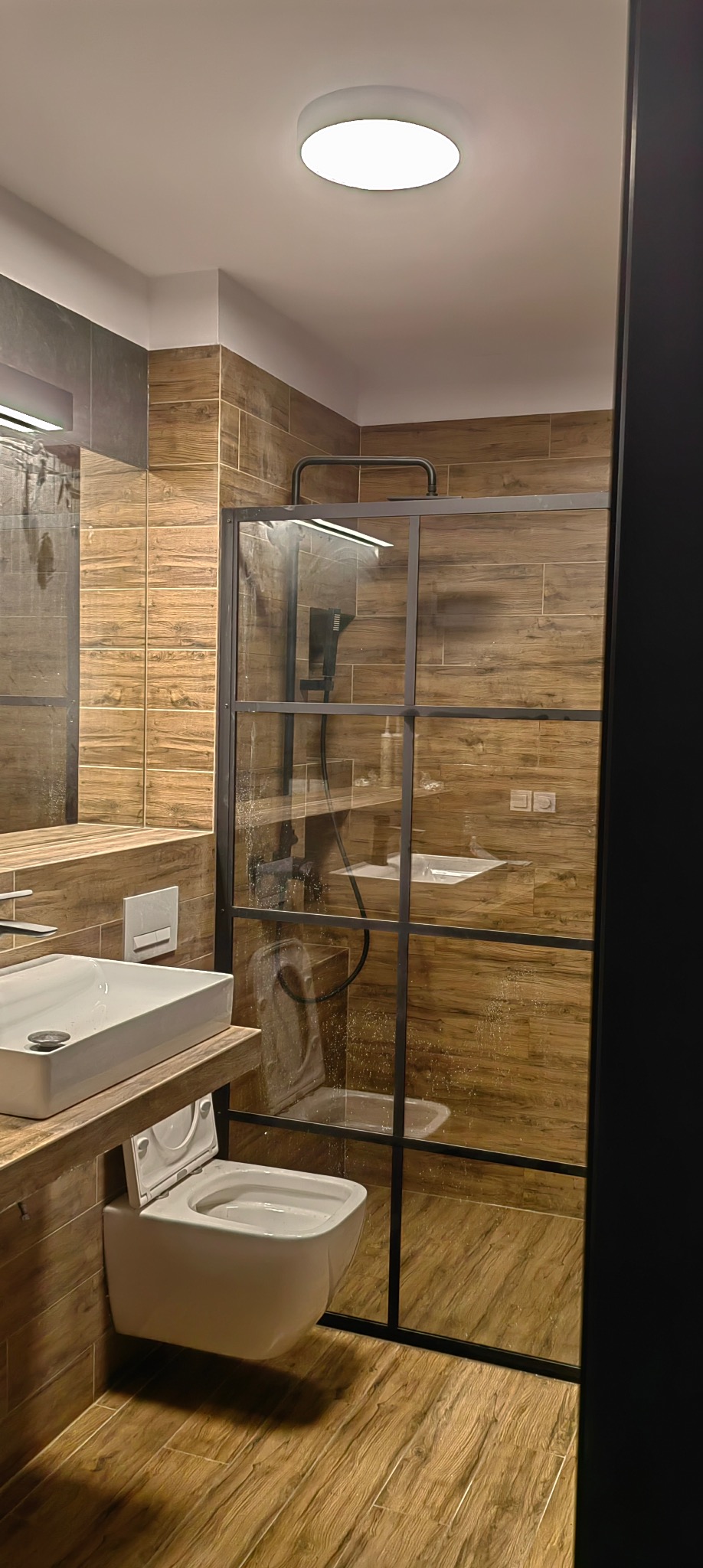 Nowoczesna łazienka z kabiną prysznicową typu walk-in, drewnopodobnymi płytkami i podwieszaną toaletą. Minimalistyczny design, ciepłe oświetlenie.