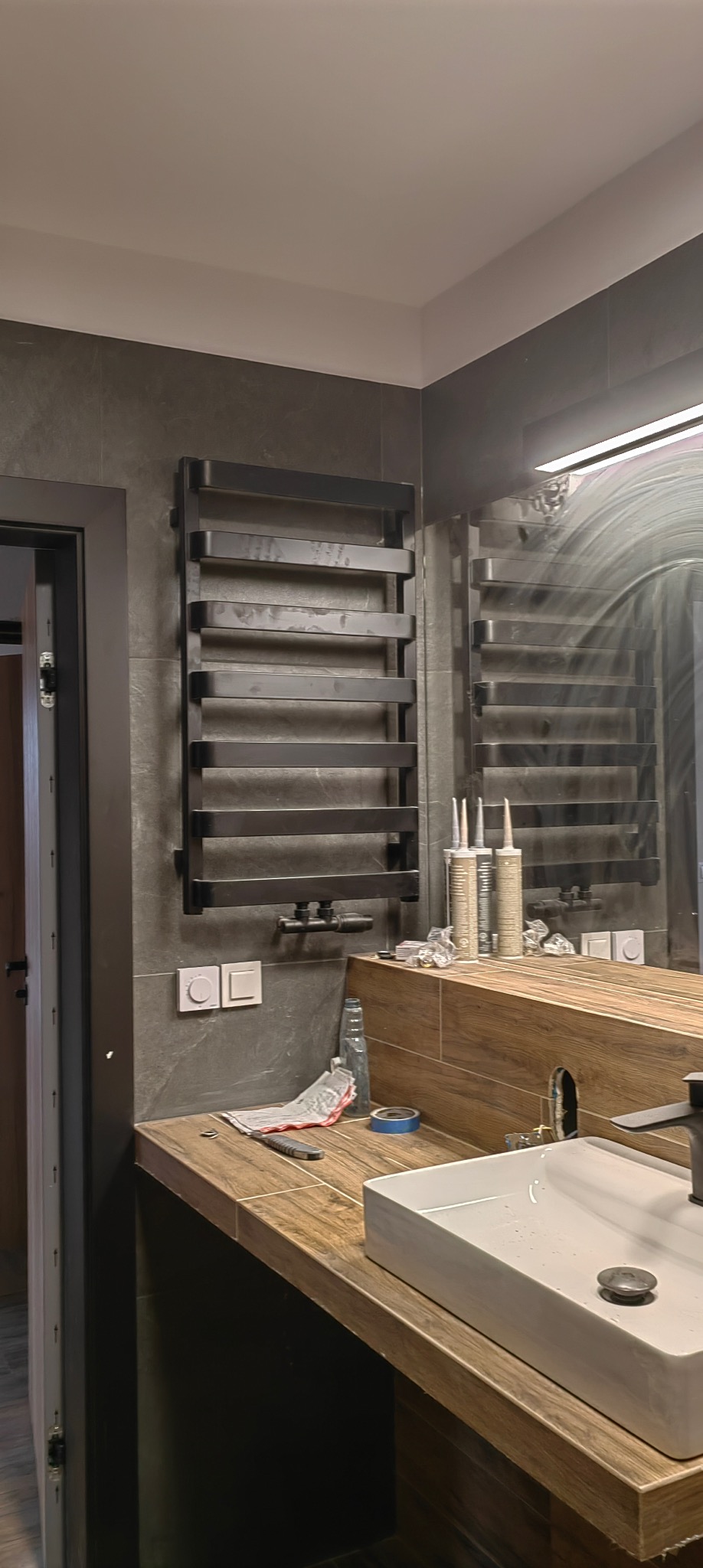 Nowoczesna łazienka z czarnym grzejnikiem drabinkowym na szarej ścianie z płytek, drewnopodobny blat z białą umywalką i widoczne narzędzia glazurnicze.