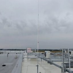 ADVOLT - Instalacja odgromowa na dachu budynku z widocznymi elementami mocującymi i przewodami. W tle pochmurne niebo i fragmenty innych instalacji.