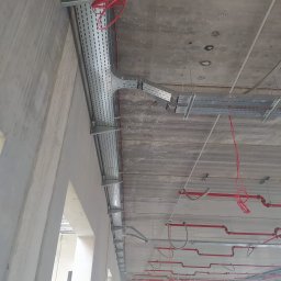 ADVOLT - Szara, perforowana trasa kablowa zamontowana pod betonowym stropem. Widoczne czerwone kable i rury. W tle fragment ściany i sylwetka osoby w kasku.