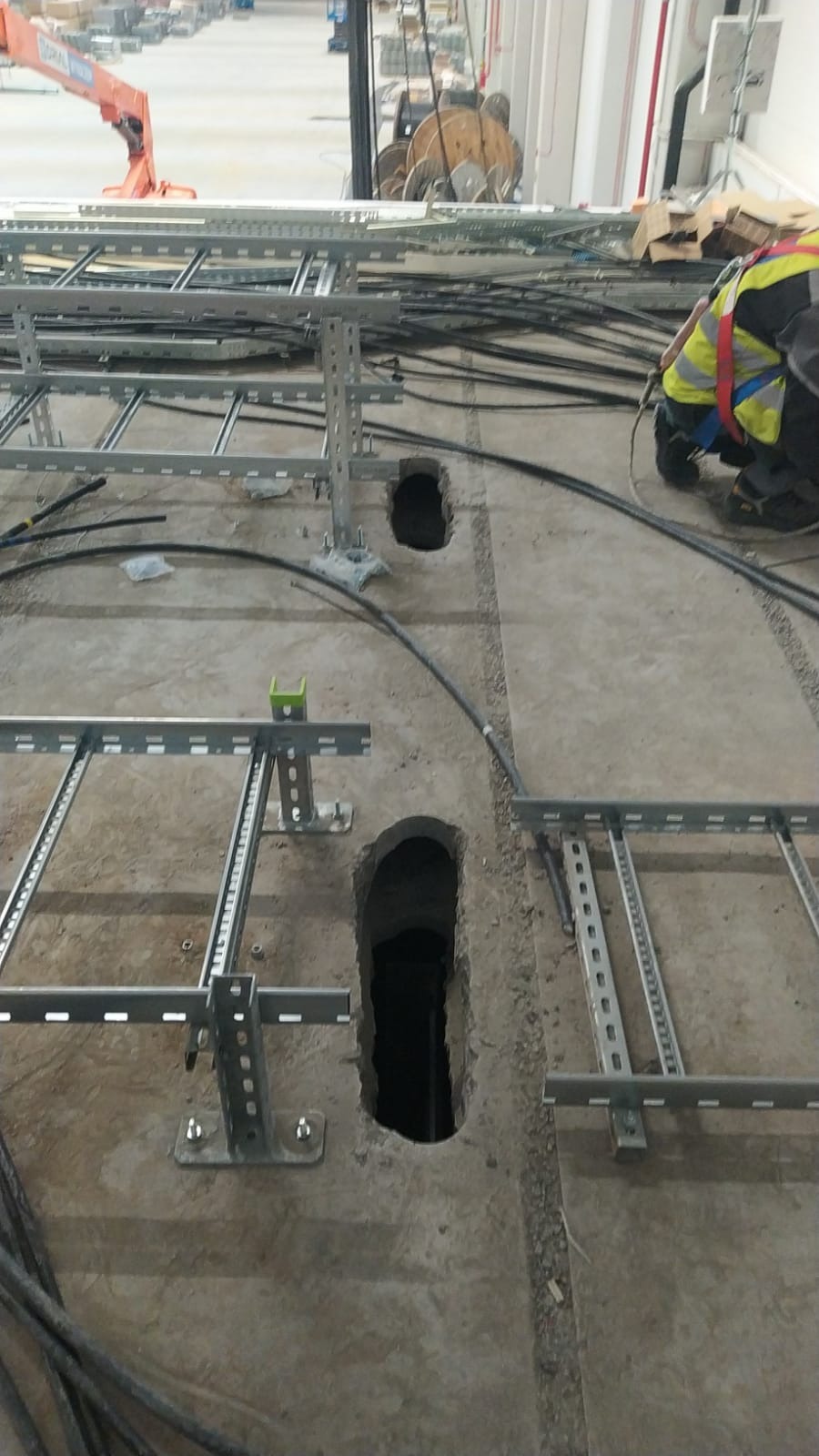 Montaż tras kablowych w podłodze z wywierconymi otworami. Widoczny pracownik w kamizelce odblaskowej. Kable ułożone wzdłuż metalowych konstrukcji.