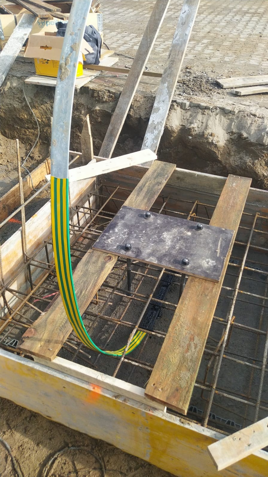 Uziemienie fundamentu z żółto-zielonym przewodem w szalunku zbrojeniowym. Metalowa płyta z nakrętkami. Widok z góry na plac budowy.