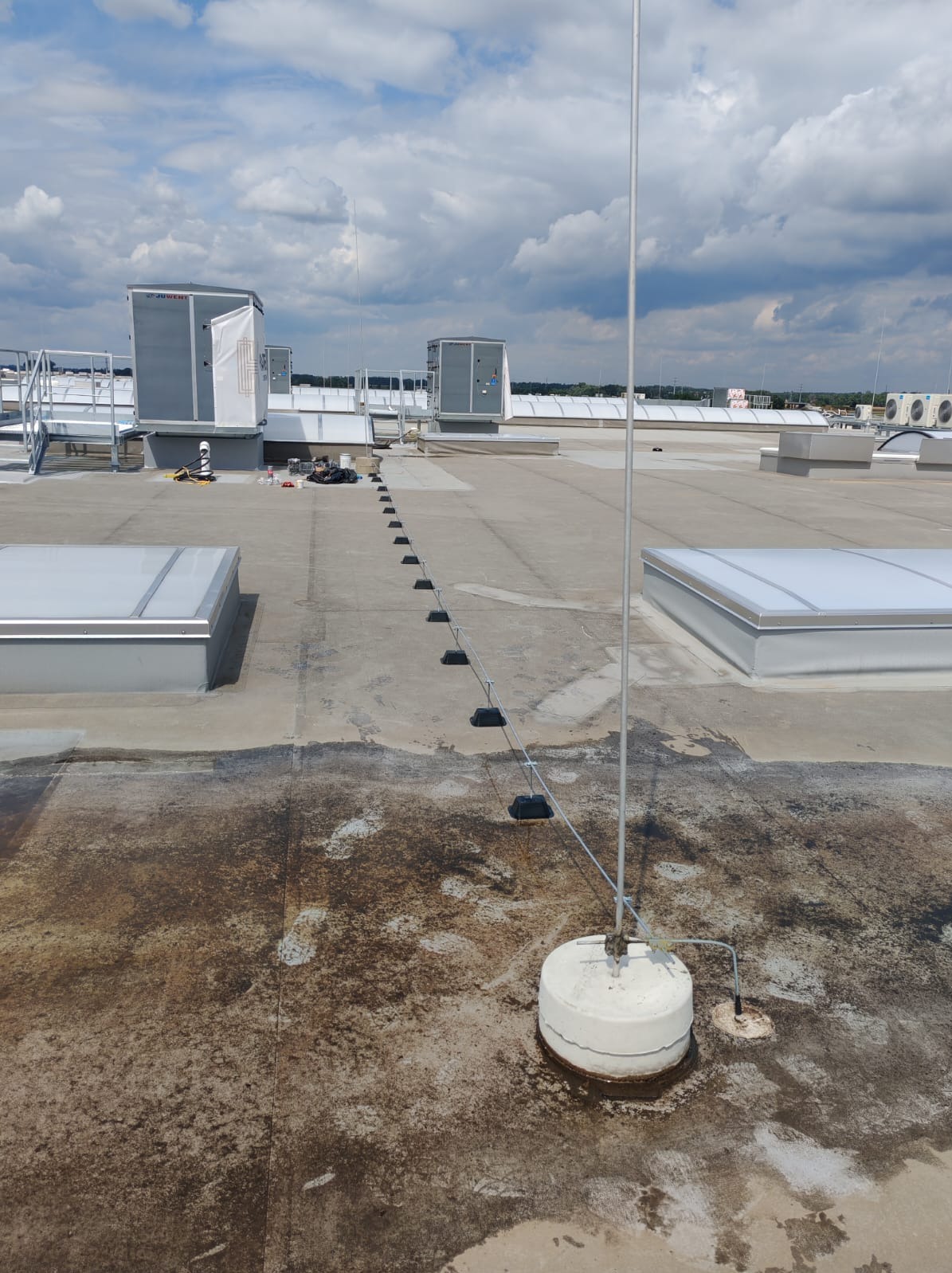 Instalacja odgromowa na dachu budynku z widocznymi elementami mocującymi i przewodami. Tło stanowią urządzenia techniczne i błękitne niebo z chmurami.