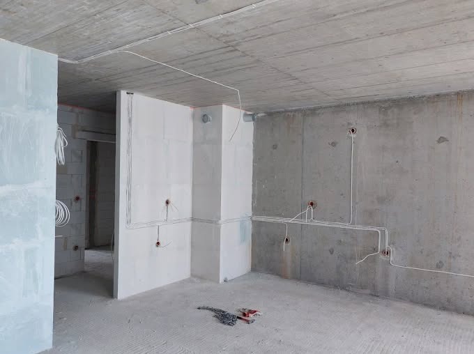 Surowy stan wnętrza z widoczną instalacją elektryczną: kable prowadzone po ścianach i suficie, przygotowane punkty pod gniazdka. Widoczne narzędzia na podłodze.