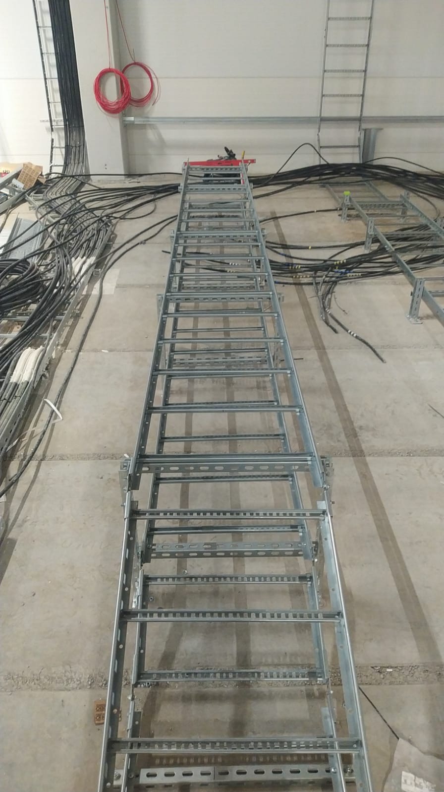 Metalowa drabinka kablowa zamontowana w hali, z widocznymi kablami ułożonymi na ścianach i suficie. Widok z dołu, perspektywa zbieżna. Dwa zwisające kable w kolorze czerwonym.