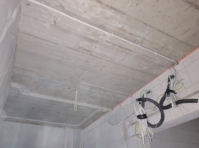 Instalacja elektryczna w surowym wnętrzu: kable prowadzone po suficie i ścianach, zabezpieczone uchwytami. Widoczne przewody w peszlach i korytkach.