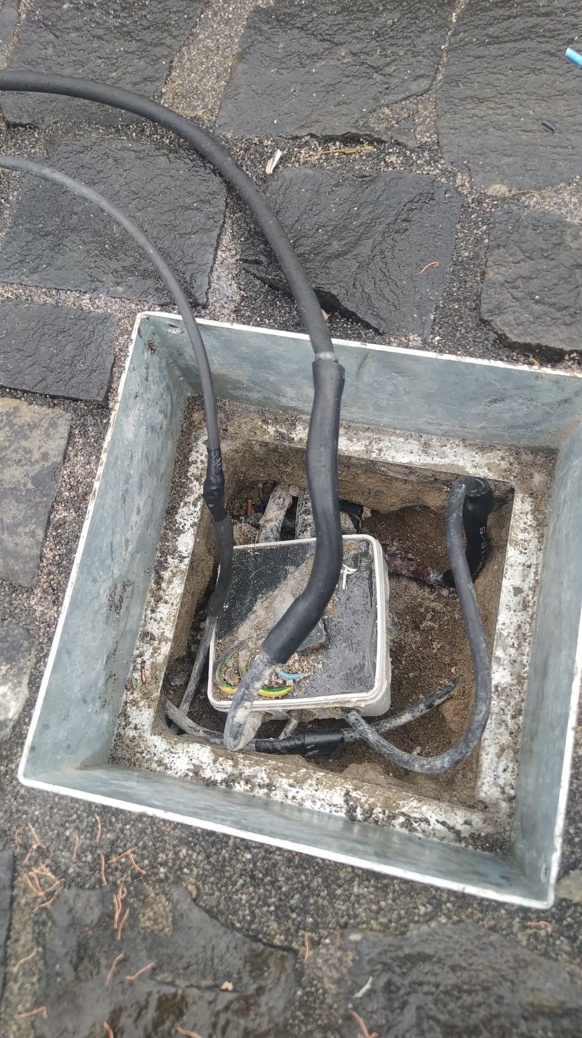Otwarta, metalowa skrzynka rewizyjna z kablami elektrycznymi na zewnątrz, umieszczona na mokrej, kamiennej nawierzchni. Widoczne złącza kablowe i elementy instalacji.