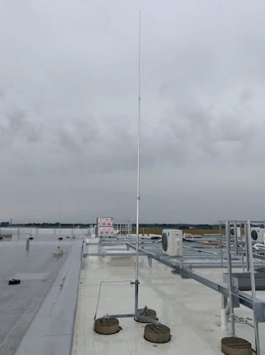 Instalacja odgromowa na dachu budynku z widocznymi elementami mocującymi i przewodami. W tle pochmurne niebo i fragmenty innych instalacji.