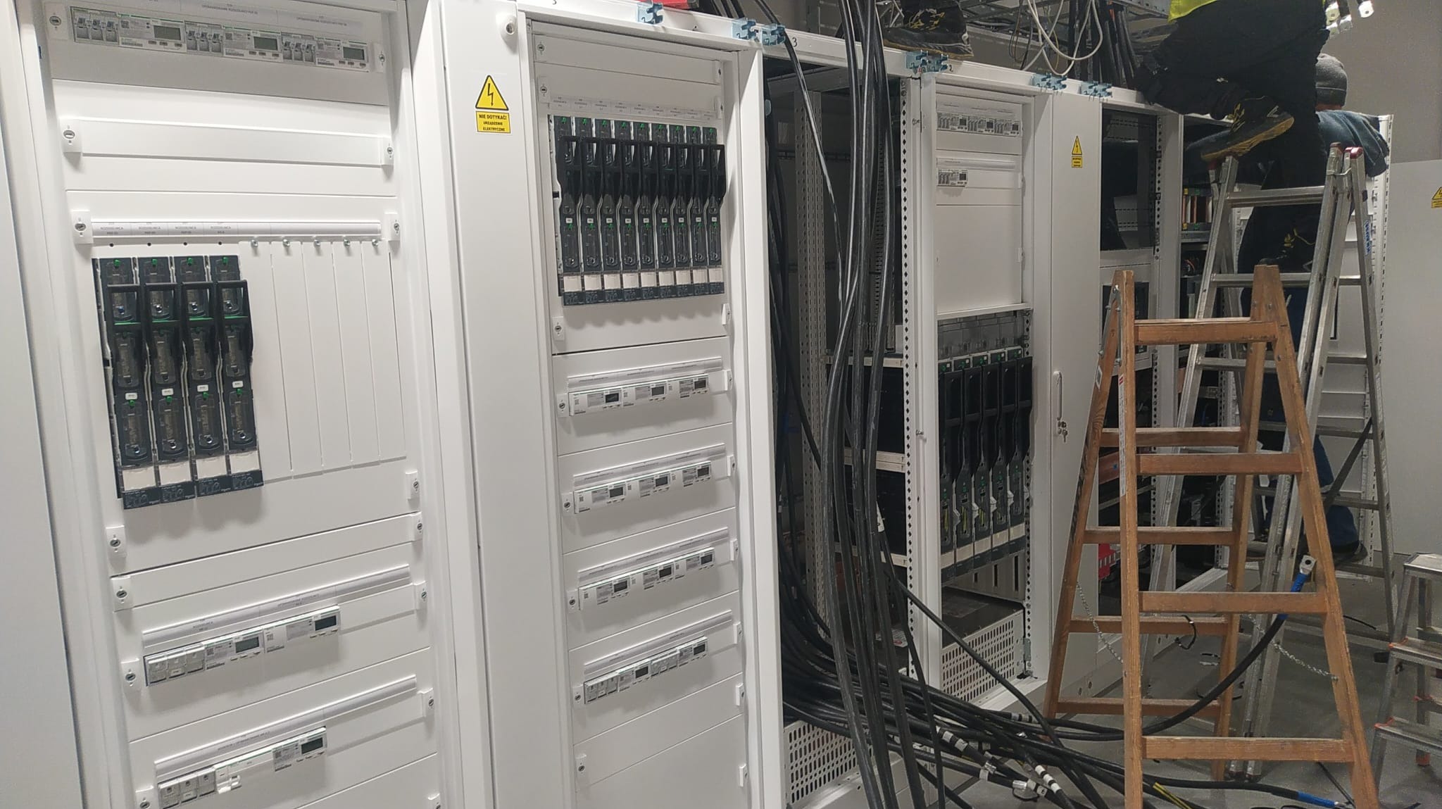Rozdzielnice elektryczne w białych obudowach, widoczne kable i drabiny, sugerujące prace instalacyjne w dużej serwerowni lub obiekcie przemysłowym.
