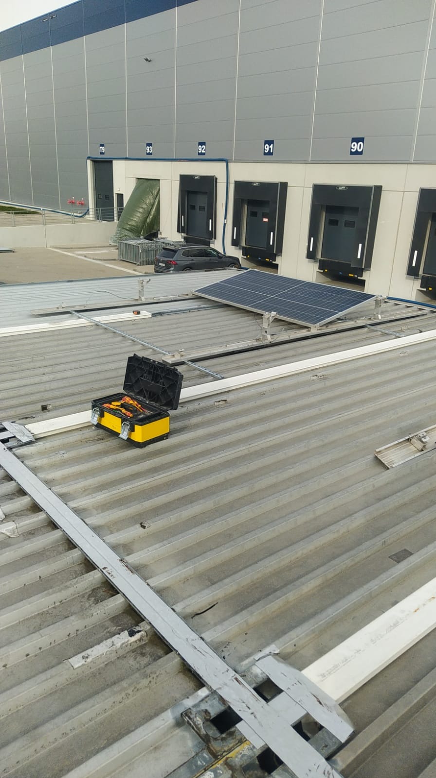 Montaż paneli słonecznych na dachu hali magazynowej. Otwarta skrzynka narzędziowa z narzędziami elektrycznymi obok panelu. Widoczne elementy mocujące i konstrukcja wsporcza.