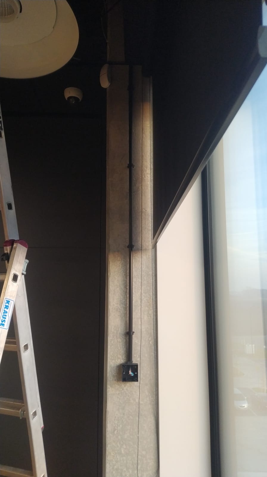 Zamknięta puszka elektryczna z poprowadzonym kablem na betonowym słupie, widoczna drabina Krause, fragment okna i lampy sufitowej.