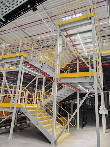 Wielopoziomowa konstrukcja stalowa z żółtymi akcentami w hali magazynowej. Schody z antypoślizgowymi nakładkami. Widoczne instalacje pod sufitem.