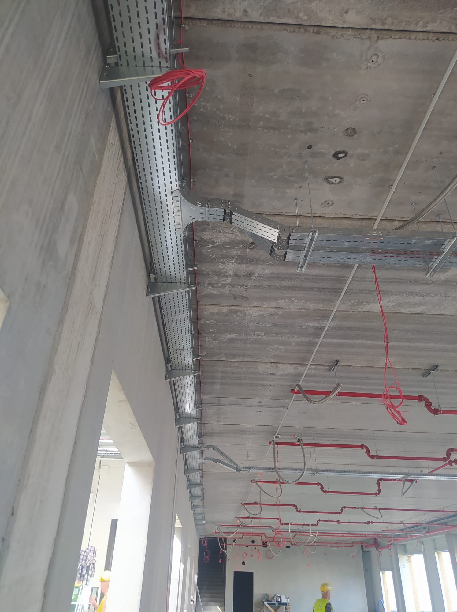 Szara, perforowana trasa kablowa zamontowana pod betonowym stropem. Widoczne czerwone kable i rury. W tle fragment ściany i sylwetka osoby w kasku.