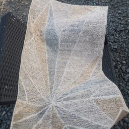 Clinex Paulina Ciak - Wzorzysty, jasny dywan z geometrycznym motywem, częściowo zwinięty, leży na czarnym, plecionym krześle na tle szarego żwiru. Widoczny fragment buta.