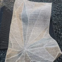 Wzorzysty, jasny dywan z geometrycznym motywem, częściowo zwinięty, leży na czarnym, plecionym krześle na tle szarego żwiru. Widoczny fragment buta.