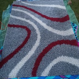 Clinex Paulina Ciak - Wzór dywanu z szarym tłem i czerwono-białymi, zakrzywionymi pasami, rozłożony na trawie, sugerujący pranie lub suszenie na zewnątrz.