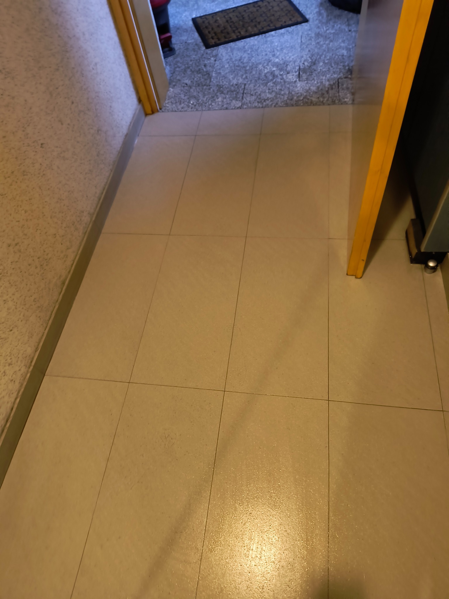 Beżowe płytki podłogowe w korytarzu, widok z góry, z fragmentem drzwi po prawej i wejściem do innego pomieszczenia z wycieraczką w tle.