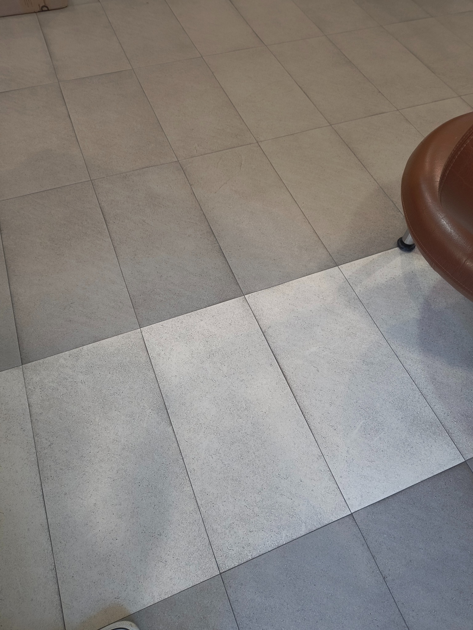 Ułożone, kwadratowe płytki podłogowe w odcieniach szarości i beżu, z fragmentem brązowego fotela w prawym górnym rogu. Widoczny fragment buta w lewym dolnym rogu.