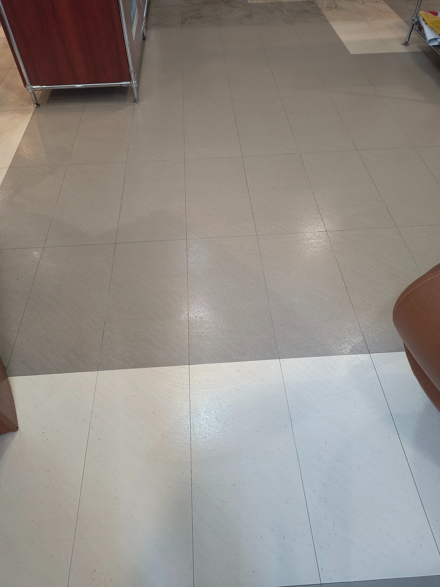 Wypolerowana podłoga z płytek w odcieniach szarości i bieli, z fragmentem mebla w rogu. Widoczne delikatne refleksy światła na powierzchni płytek.