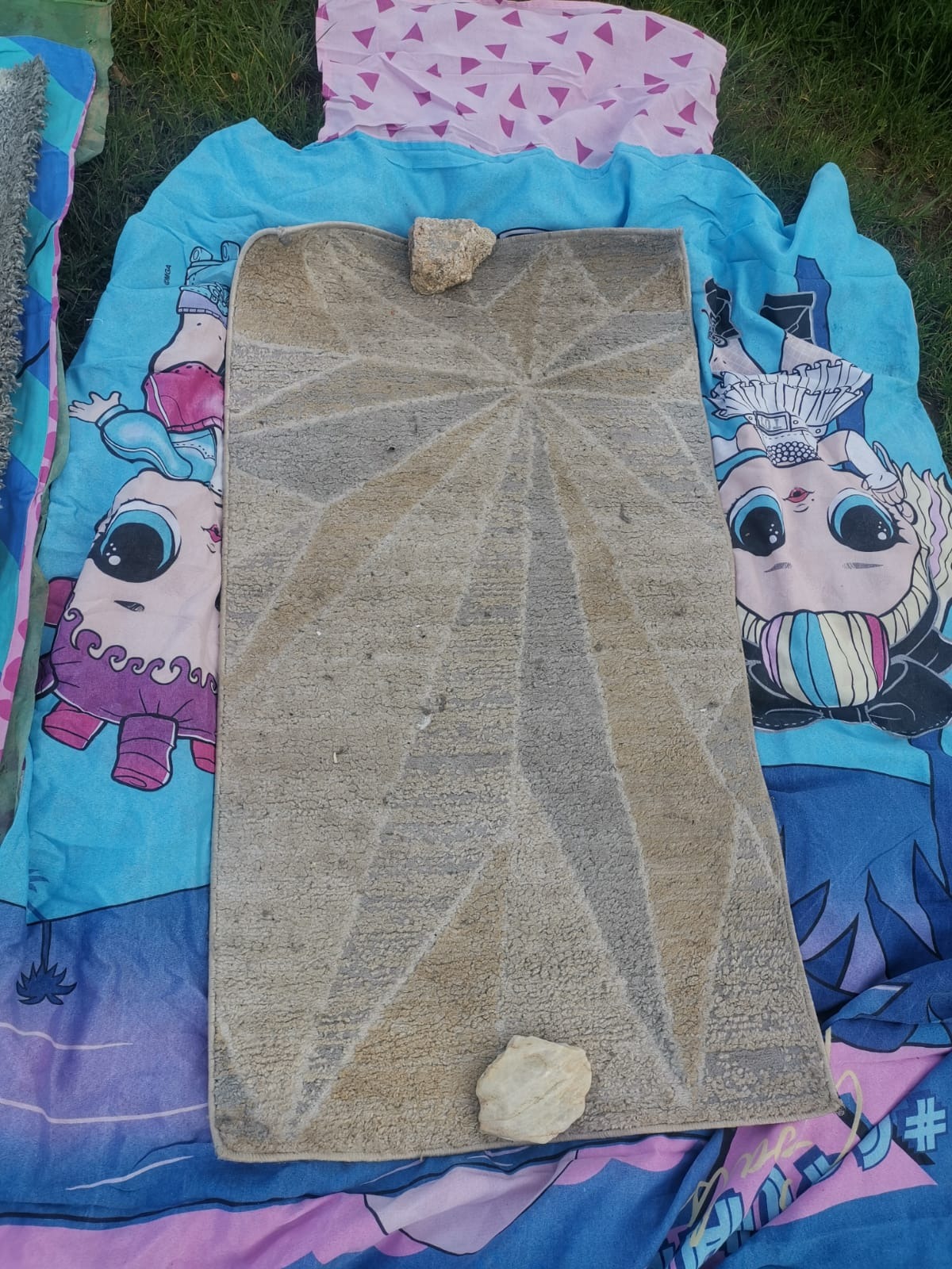 Brudny, beżowy dywan z geometrycznym wzorem leży na kolorowej pościeli z postaciami z bajek. Na dywanie dwa kamienie. Widok z góry.