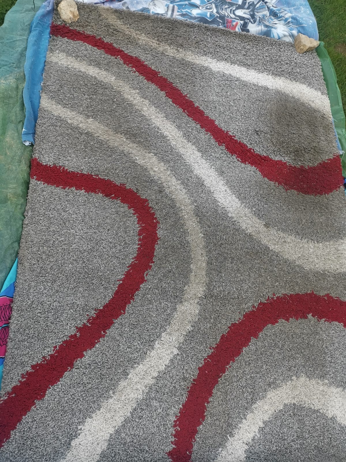 Szary dywan z czerwonymi i białymi wzorami falistymi, leżący na zewnątrz, przytrzymywany kamieniami na rogach. Widoczne ślady użytkowania.