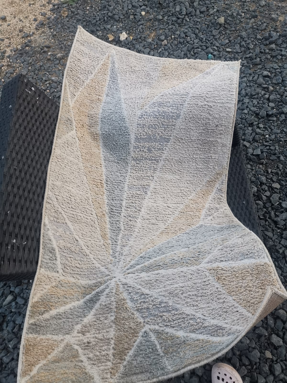 Wzorzysty, jasny dywan z geometrycznym motywem, częściowo zwinięty, leży na czarnym, plecionym krześle na tle szarego żwiru. Widoczny fragment buta.