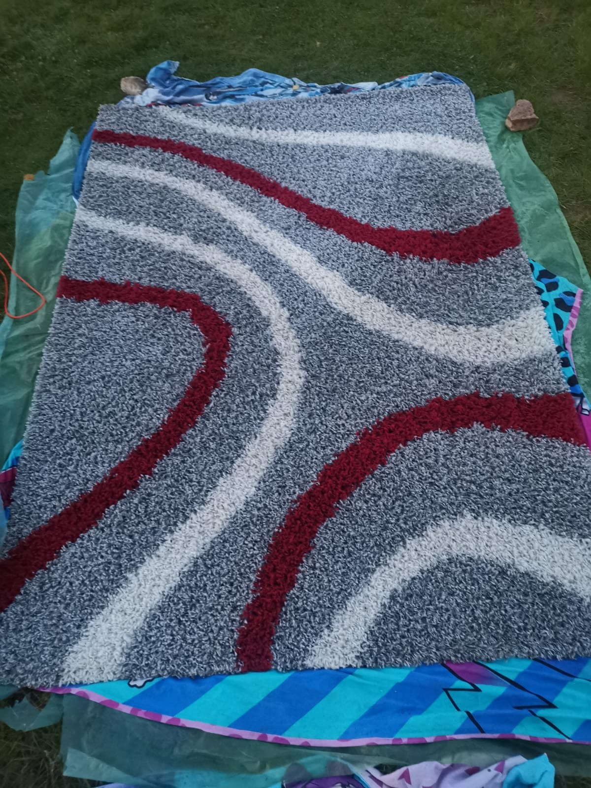 Wzór dywanu z szarym tłem i czerwono-białymi, zakrzywionymi pasami, rozłożony na trawie, sugerujący pranie lub suszenie na zewnątrz.