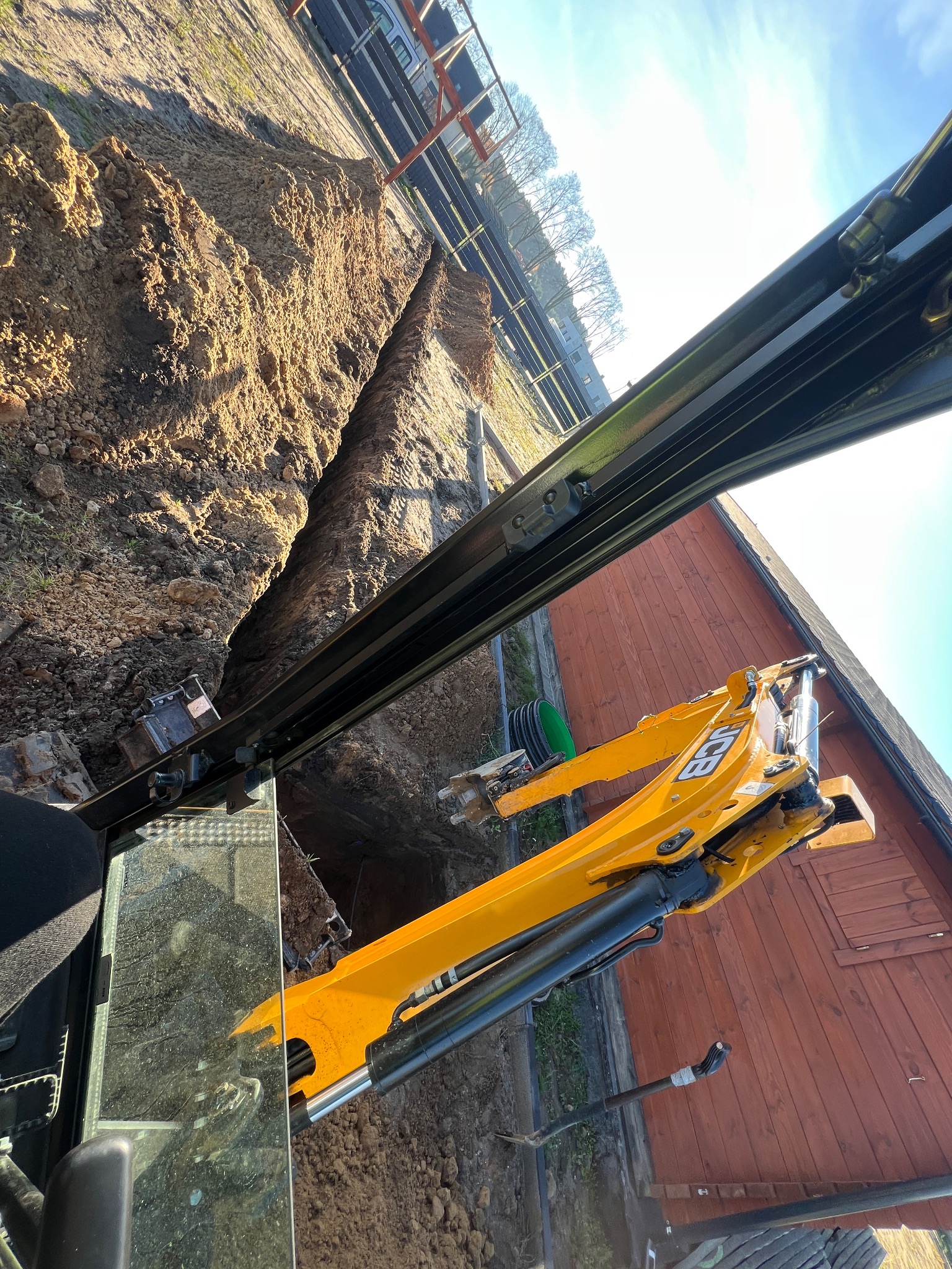 Żółta koparka JCB w trakcie wykopu wąskiego rowu w ziemi, widok z kabiny operatora, obok drewniany budynek i ogrodzenie. Słoneczny dzień.