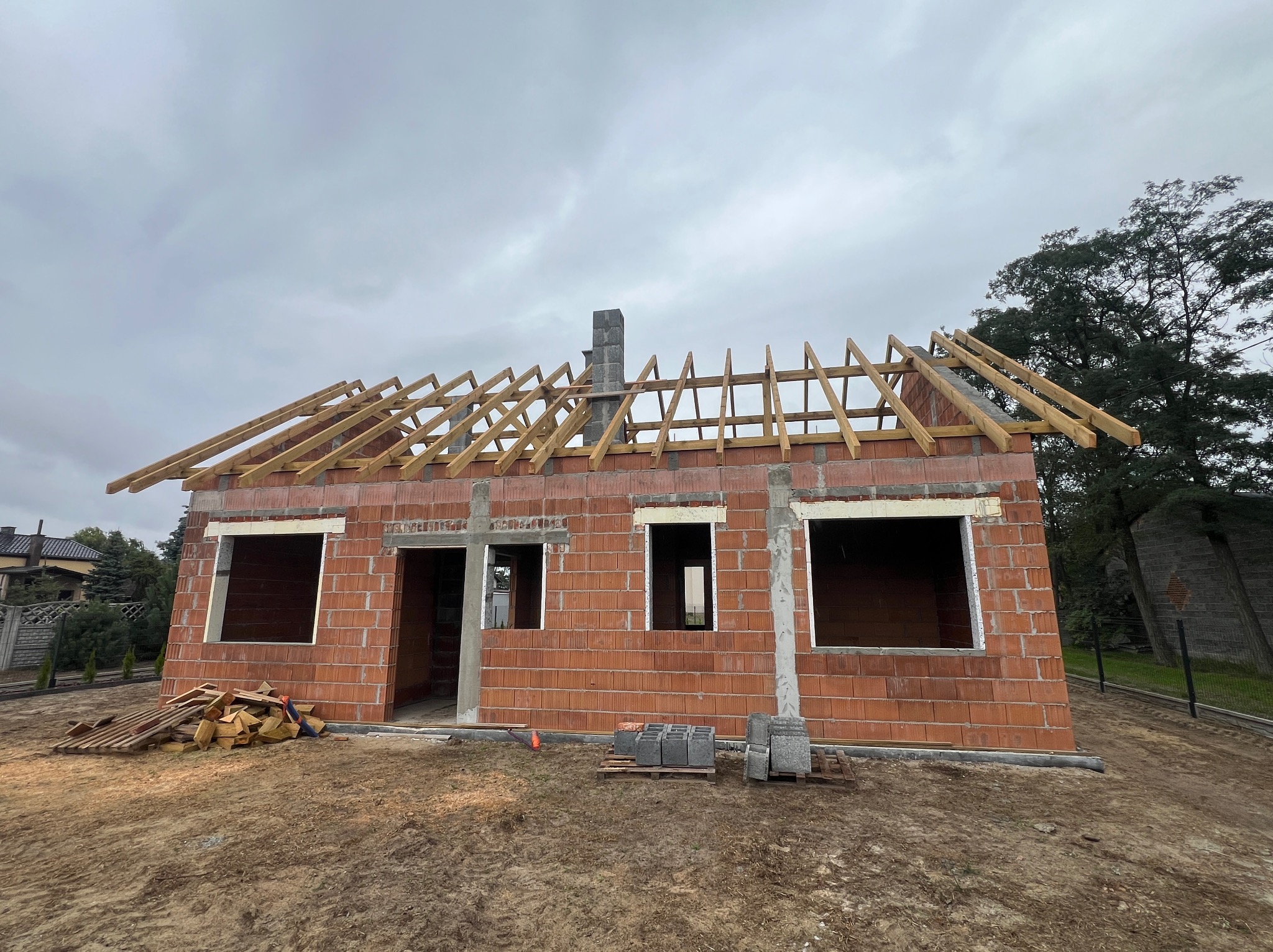 Budowa domu z czerwonej cegły: ściany wzniesione, widoczne otwory okienne i drzwiowe, drewniana konstrukcja dachu w trakcie montażu, komin. Na ziemi leżą cegły i drewniane elementy.