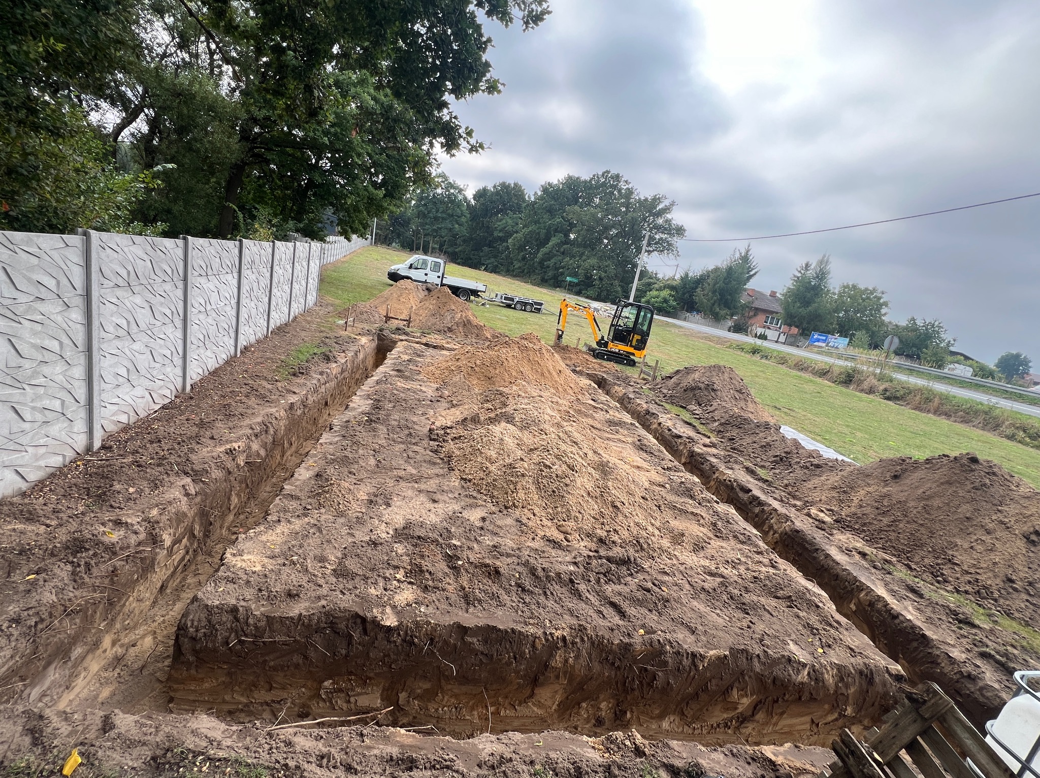 Wykopane rowy pod fundamenty z mini koparką w tle, widok z góry. Obok betonowe ogrodzenie panelowe i biały samochód dostawczy na zielonym terenie.