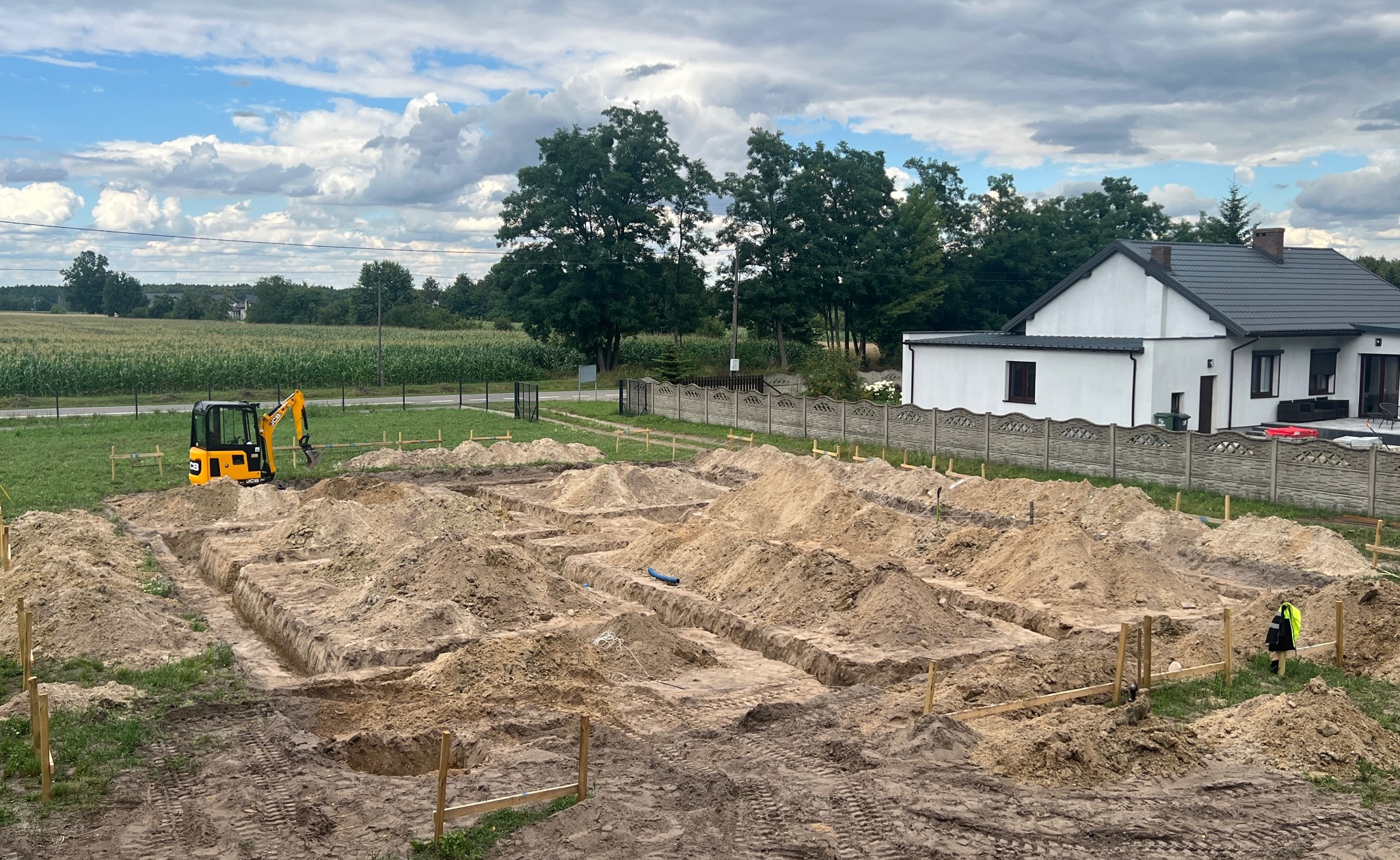 Wykopane rowy pod fundamenty domu jednorodzinnego z żółtą mini koparką JCB w tle, widok z góry na plac budowy w słoneczny dzień.