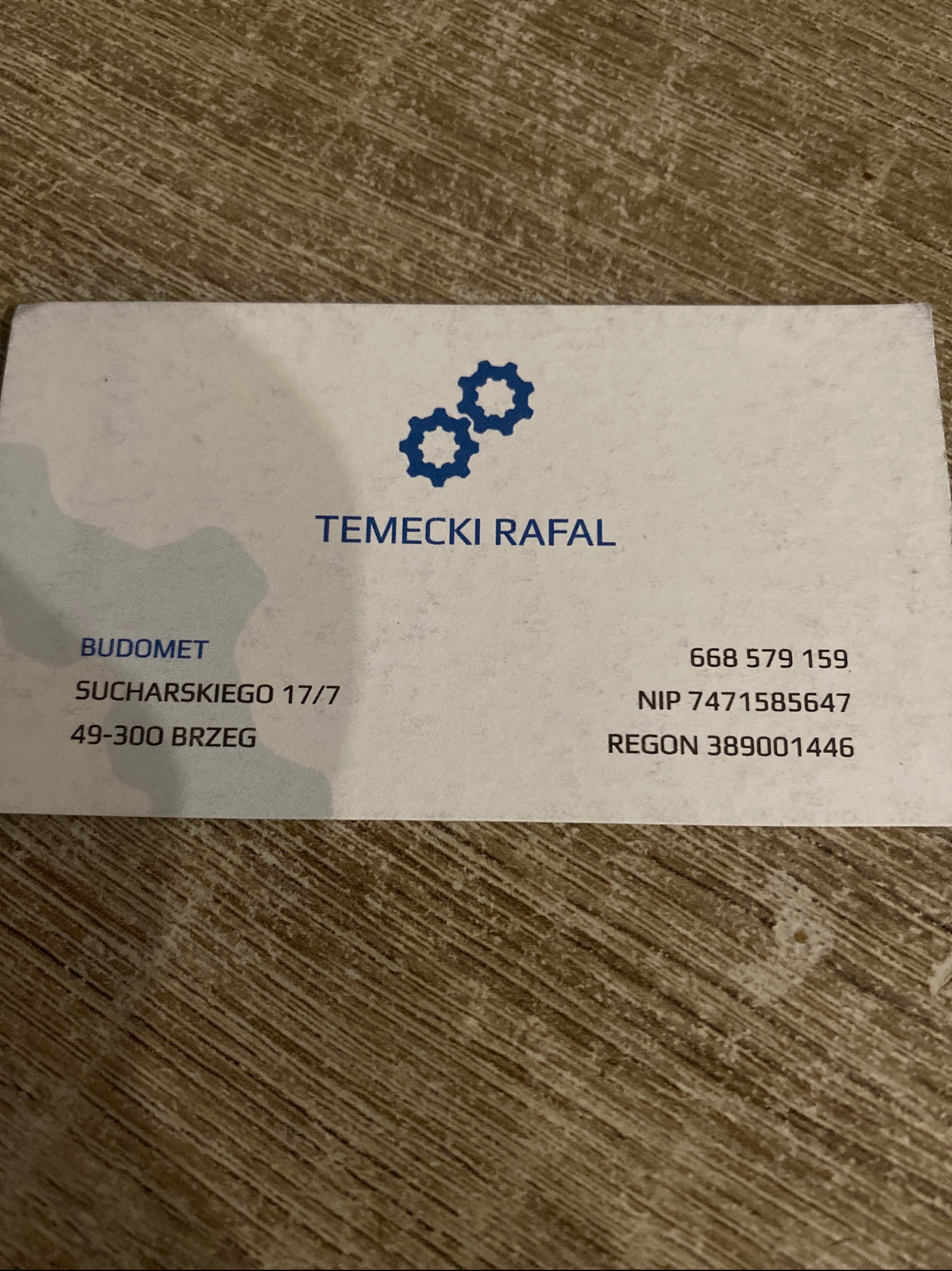 Wizytówka firmy BUDOMET Temecki Rafał z adresem Sucharskiego 17/7, Brzeg, numerem telefonu i NIP, na tle drewnopodobnej powierzchni.