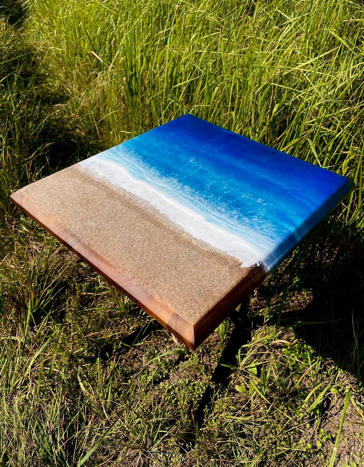 Kwadratowy stolik kawowy z blatem imitującym plażę i morze, wykonany z żywicy epoksydowej i drewna, ustawiony na trawie w słoneczny dzień.