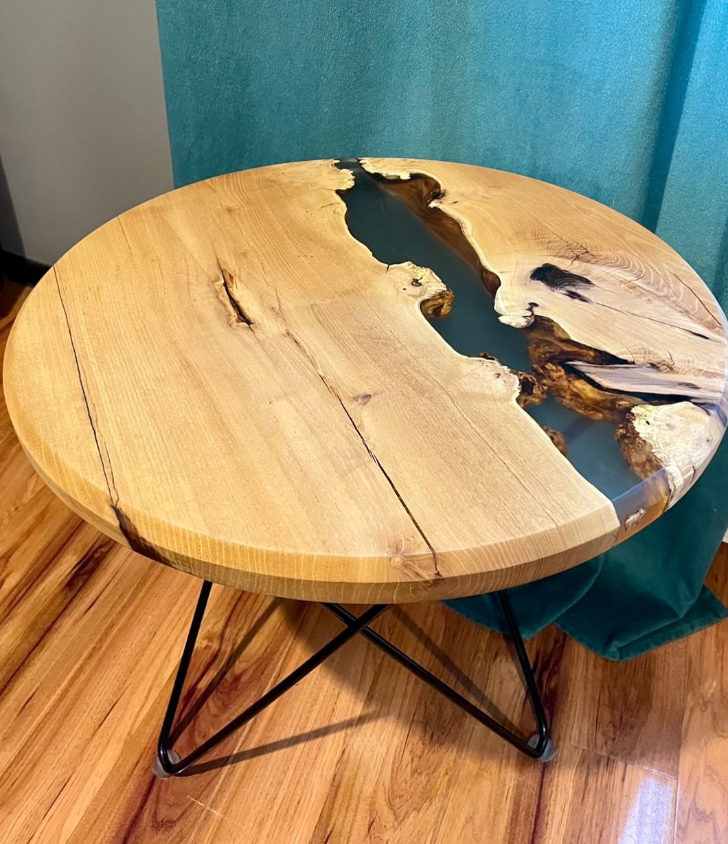 Okrągły stolik kawowy z blatem z jasnego drewna z żywicą epoksydową w kolorze morskim, osadzony na czarnych, metalowych nogach. Widoczne słoje drewna i unikalne wypełnienie żywicą.