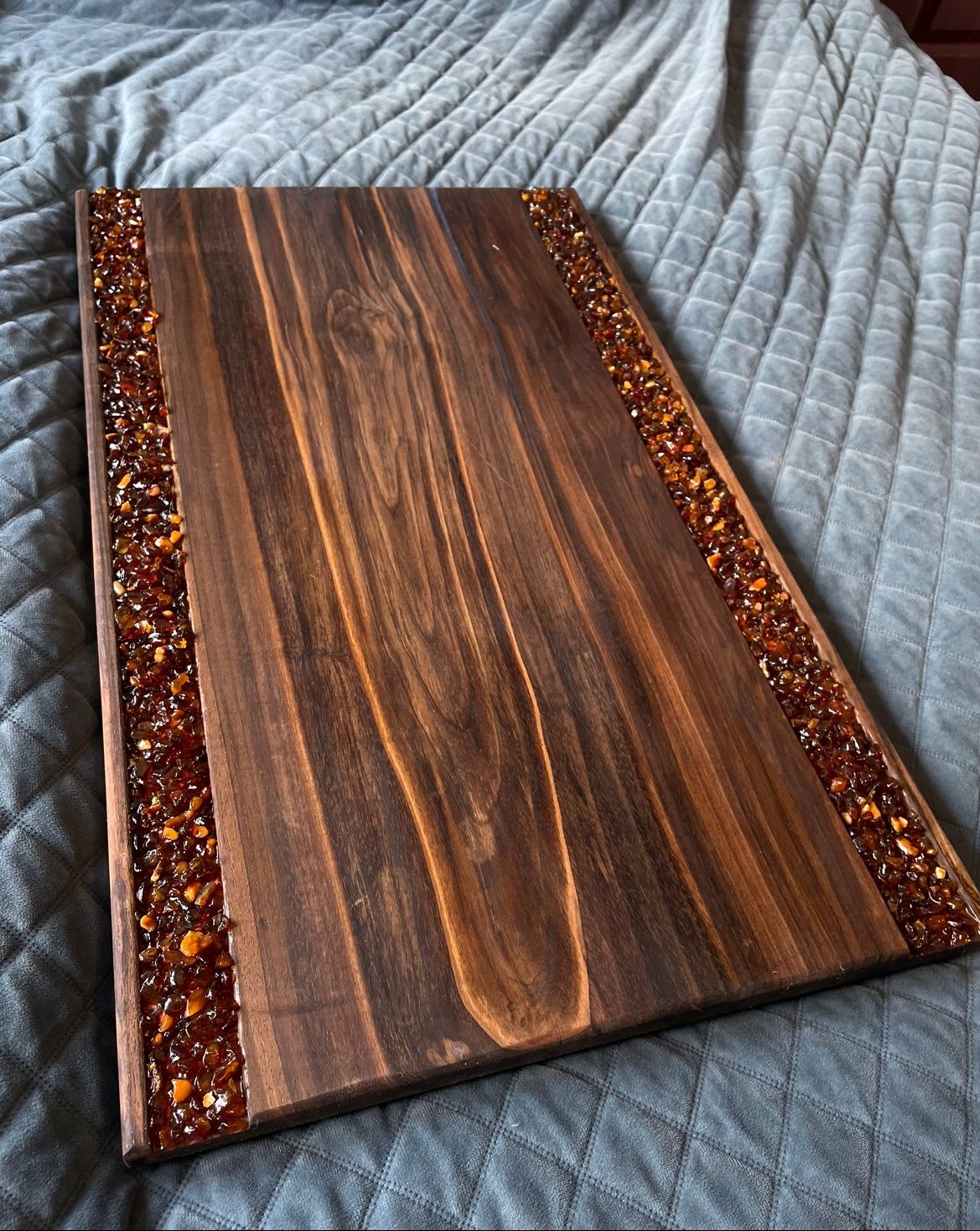 Unikatowy blat z ciemnego drewna, zdobiony po bokach bursztynową żywicą, leżący na szarym, pikowanym pledzie. Widoczne naturalne usłojenie drewna i detale żywicy.