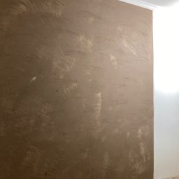 Sylwester Dobreńko - Ściana po szpachlowaniu z widoczną fakturą i śladami narzędzi, przygotowana do malowania. Fragment listwy przypodłogowej w rogu.