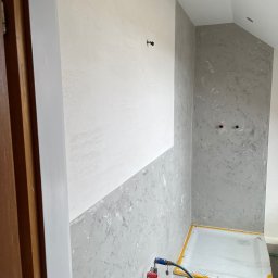 Sylwester Dobreńko - Ściana w łazience po częściowym szpachlowaniu, z widocznym podziałem na obszar biały i szary z efektem dekoracyjnym. Zabezpieczony brodzik i widoczny wąż.