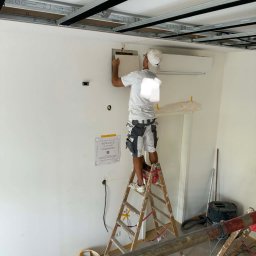 Sylwester Dobreńko - Pracownik na drabinie montuje element na ścianie pod sufitem podwieszanym. Widoczne narzędzia i akcesoria. Praca w trakcie realizacji.