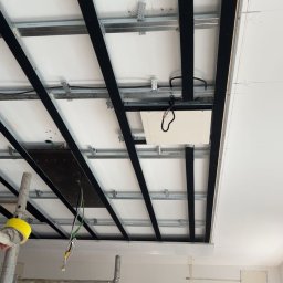 Sylwester Dobreńko - Montaż sufitu podwieszanego z widoczną konstrukcją metalową i częściowo zamontowanymi panelami. Widoczne rusztowania i przewody elektryczne.