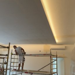 Sylwester Dobreńko - Malarz na rusztowaniu maluje sufit z oświetleniem LED. Widoczne zabezpieczone meble i narzędzia. Praca w toku, remont mieszkania.