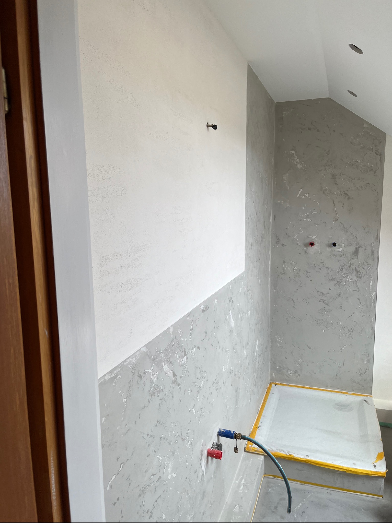 Ściana w łazience po częściowym szpachlowaniu, z widocznym podziałem na obszar biały i szary z efektem dekoracyjnym. Zabezpieczony brodzik i widoczny wąż.