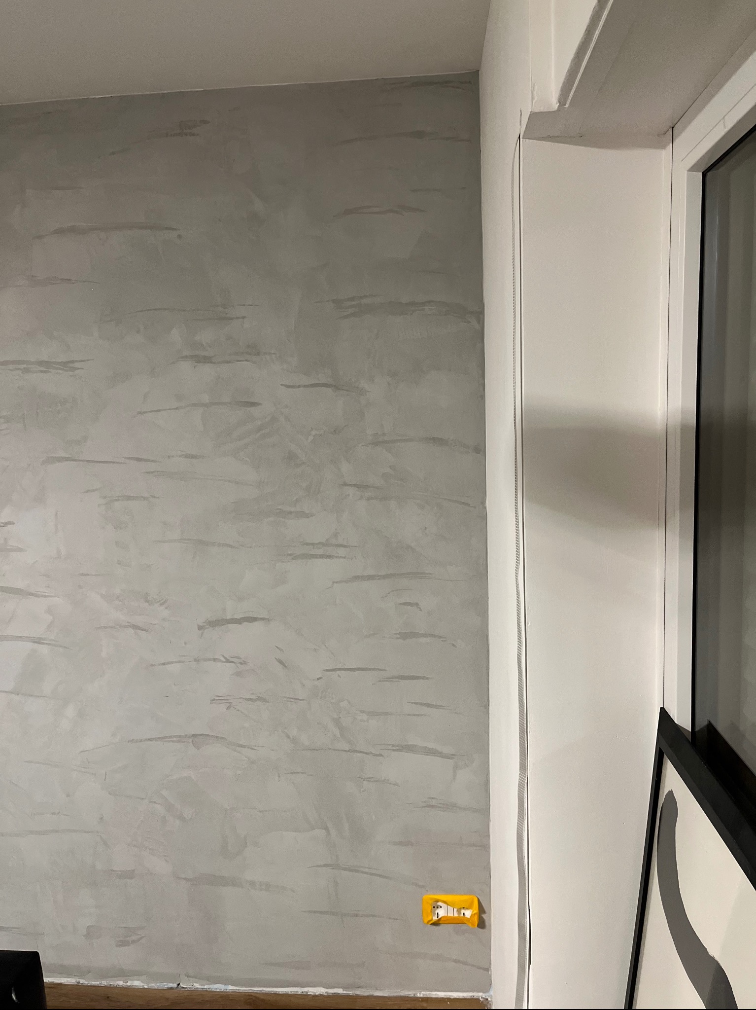 Ściana z efektem dekoracyjnym w odcieniach szarości, widoczna obok biała rama okienna i fragment obrazu w czarnej ramie. Żółte gniazdko elektryczne na ścianie.