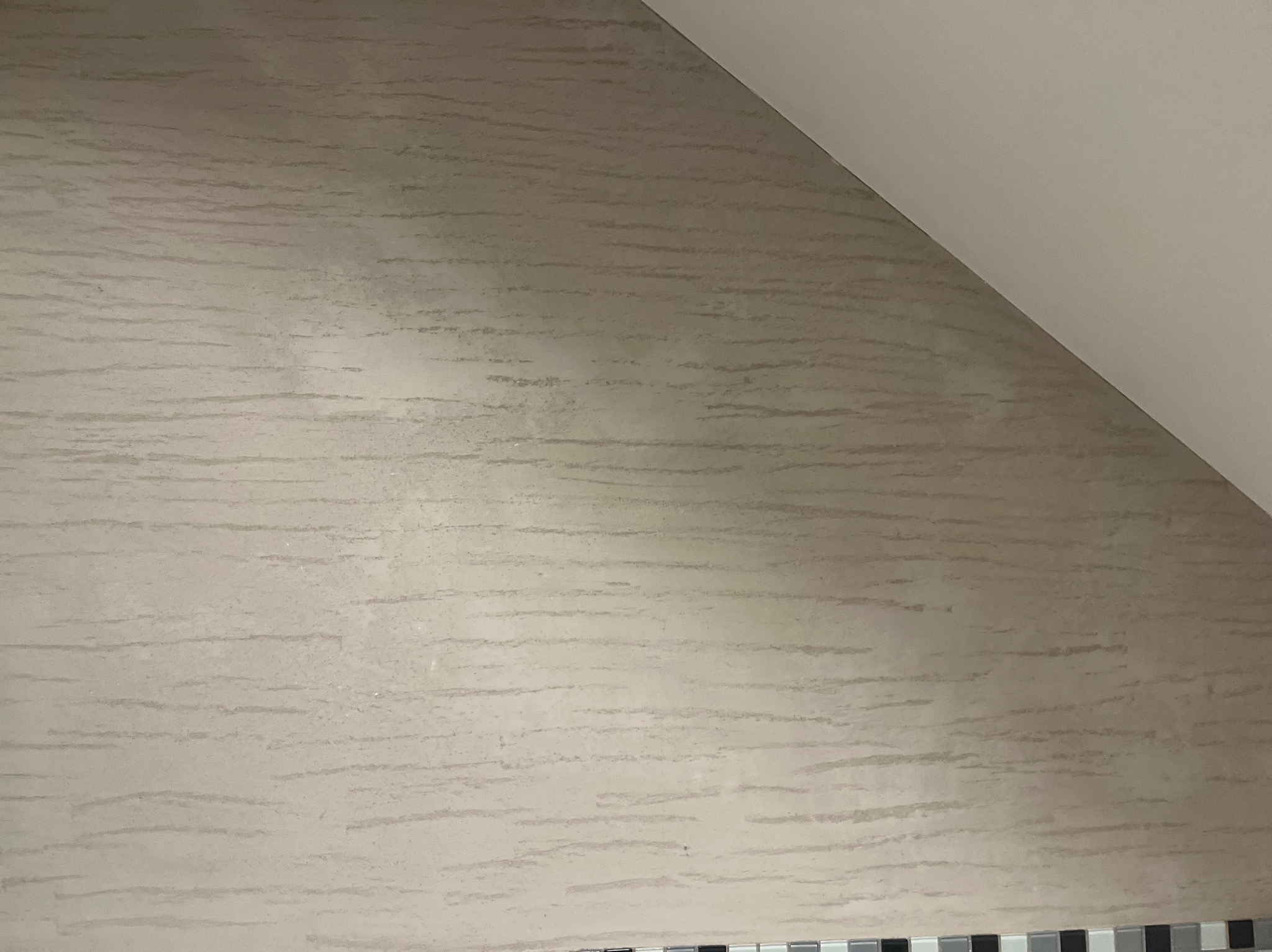 Ściana w odcieniach beżu z delikatną, poziomą fakturą przypominającą piasek, widoczna pod skosem sufitu. U dołu fragment dekoracyjnej listwy z mozaiki.