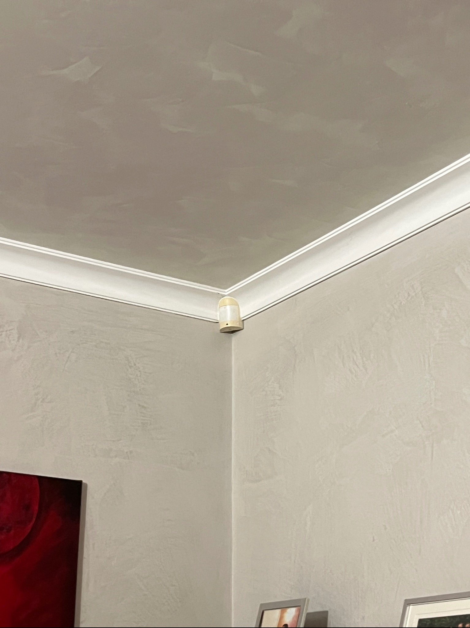 Ściana z dekoracyjną strukturą i listwą przysufitową. Widoczny narożnik pokoju, lampa sufitowa i fragment obrazu. Delikatna kolorystyka ścian.