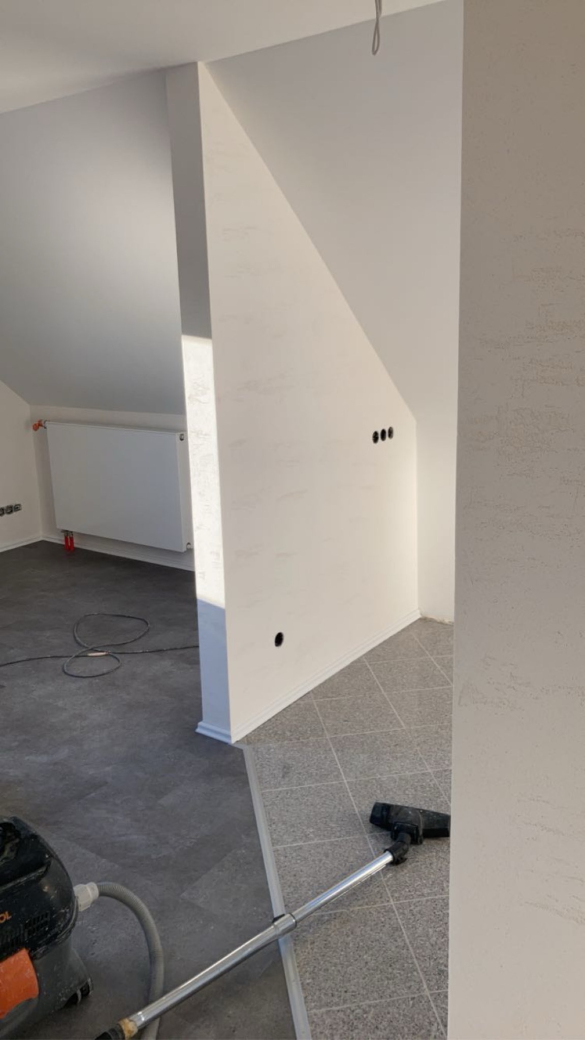 Białe ściany po remoncie w pokoju na poddaszu, widoczne gniazdka elektryczne, grzejnik i odkurzacz. Podłoga wyłożona płytkami w dwóch odcieniach szarości.