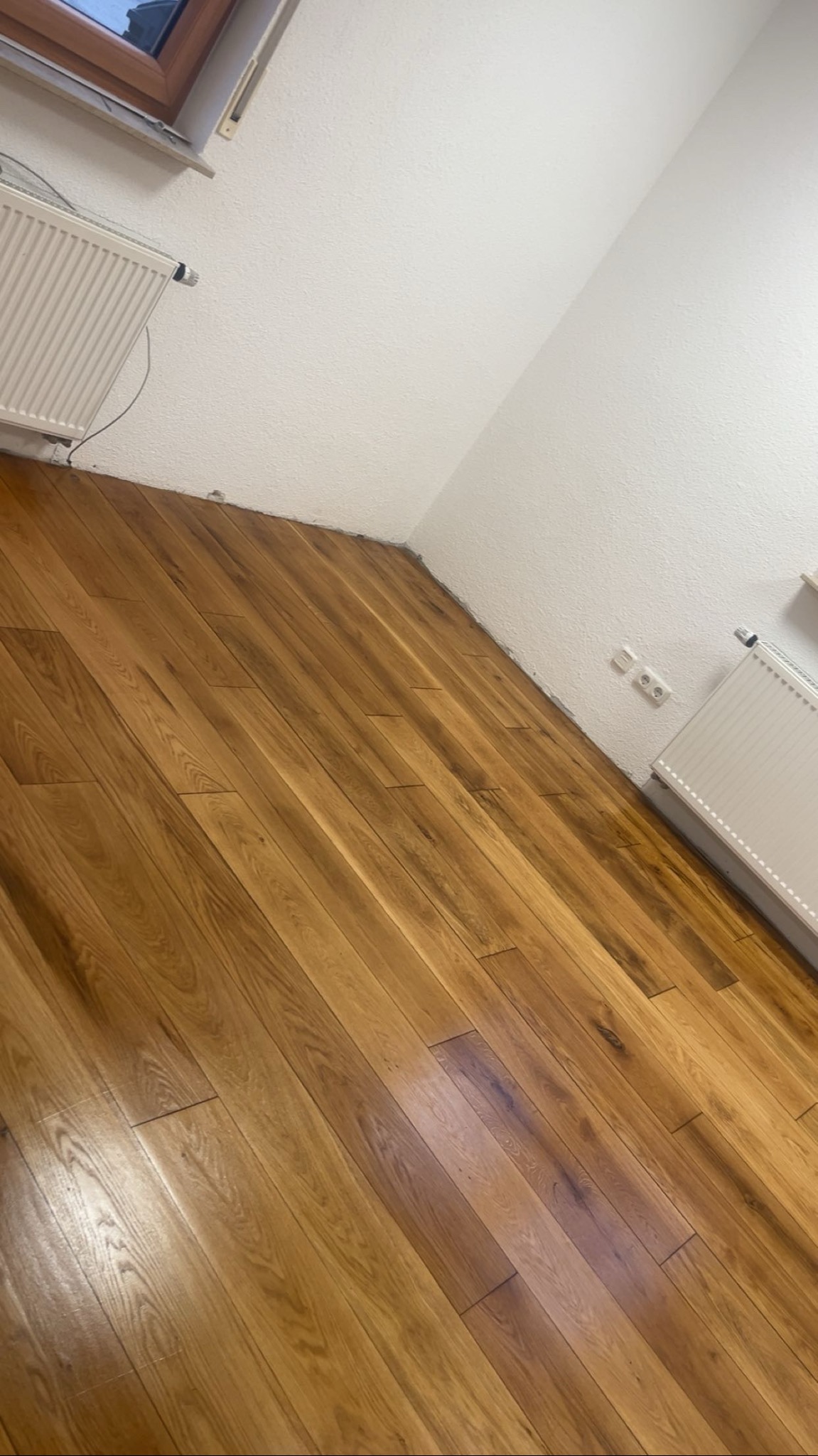 Ukończona podłoga z paneli drewnianych w rogu pokoju, widoczny grzejnik i gniazdka elektryczne. Ujęcie z dołu, podkreślające strukturę drewna.