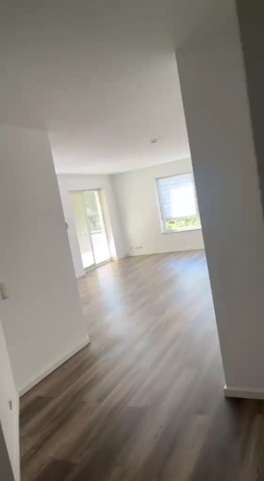 Jasne, przestronne wnętrze po remoncie: świeżo ułożone panele podłogowe w odcieniach brązu, białe ściany i widok na balkon przez otwarte drzwi.