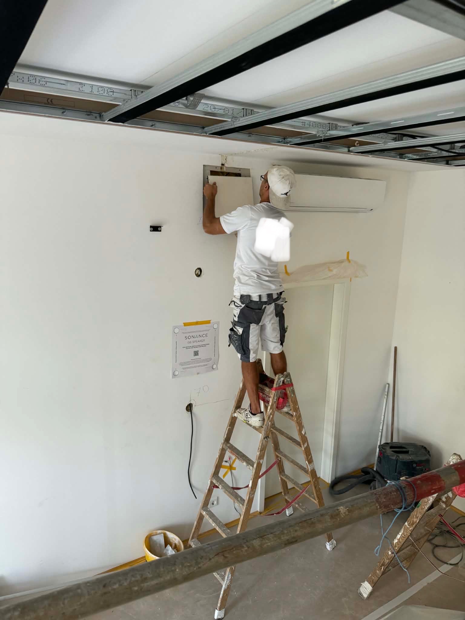 Pracownik na drabinie montuje element na ścianie pod sufitem podwieszanym. Widoczne narzędzia i akcesoria. Praca w trakcie realizacji.