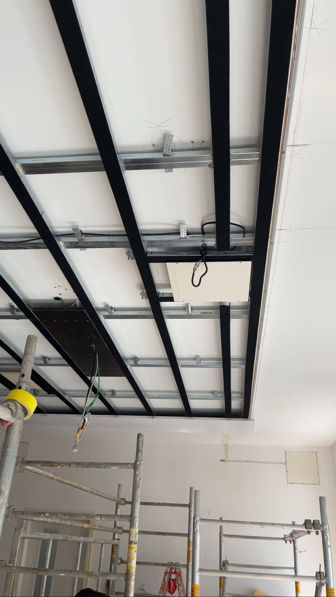 Montaż sufitu podwieszanego z widoczną konstrukcją metalową i częściowo zamontowanymi panelami. Widoczne rusztowania i przewody elektryczne.